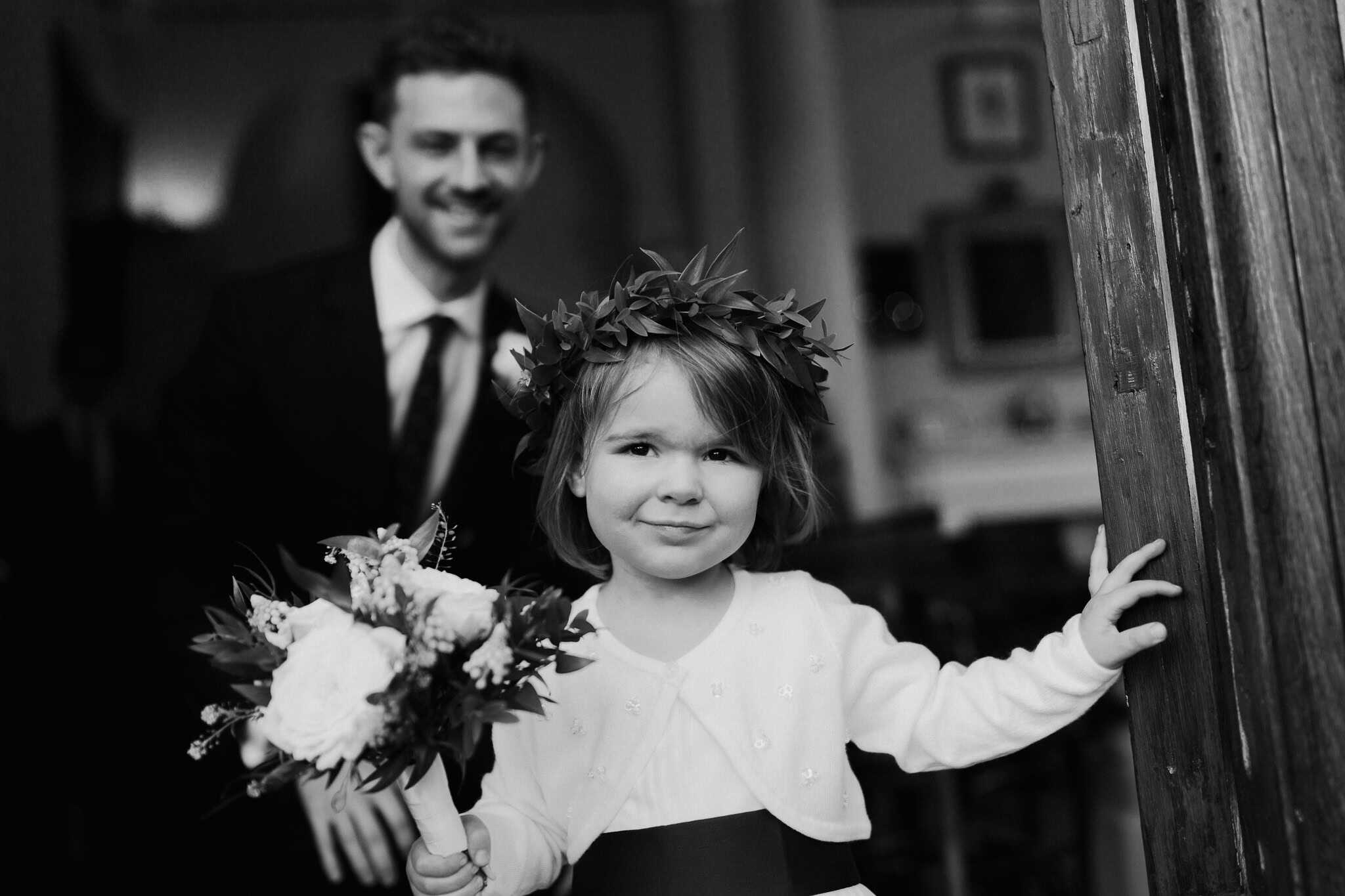 Black and white portrait of a smiling flower girl holding a bouquet at Woodstock Town Hall, with the groom in the background. A heartwarming and joyful wedding moment beautifully captured.