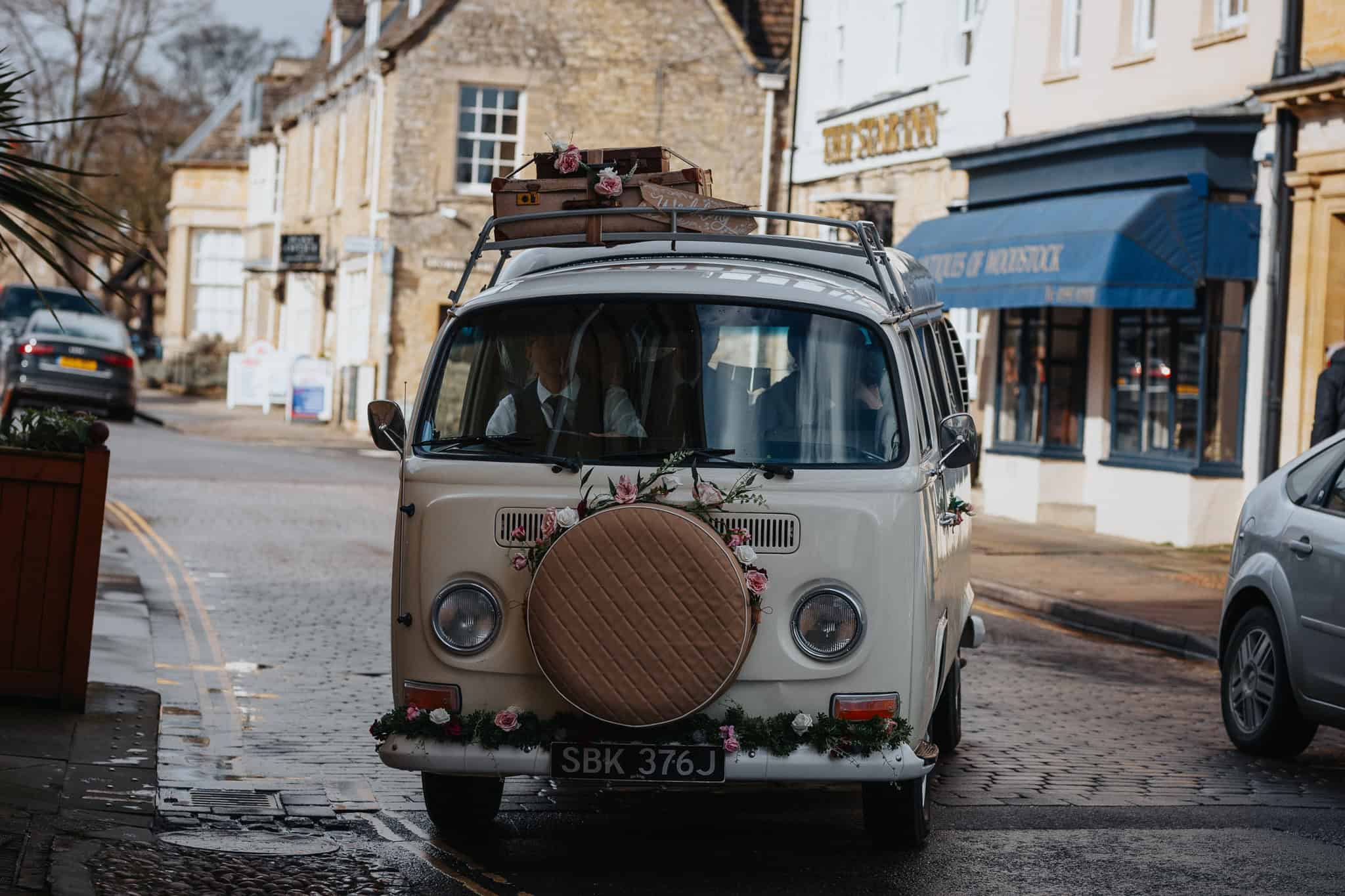 A beautifully decorated vintage VW camper van, adorned with flowers and suitcases, arriving in Woodstock, Oxfordshire, for a wedding celebration. A charming and stylish wedding transport choice.
