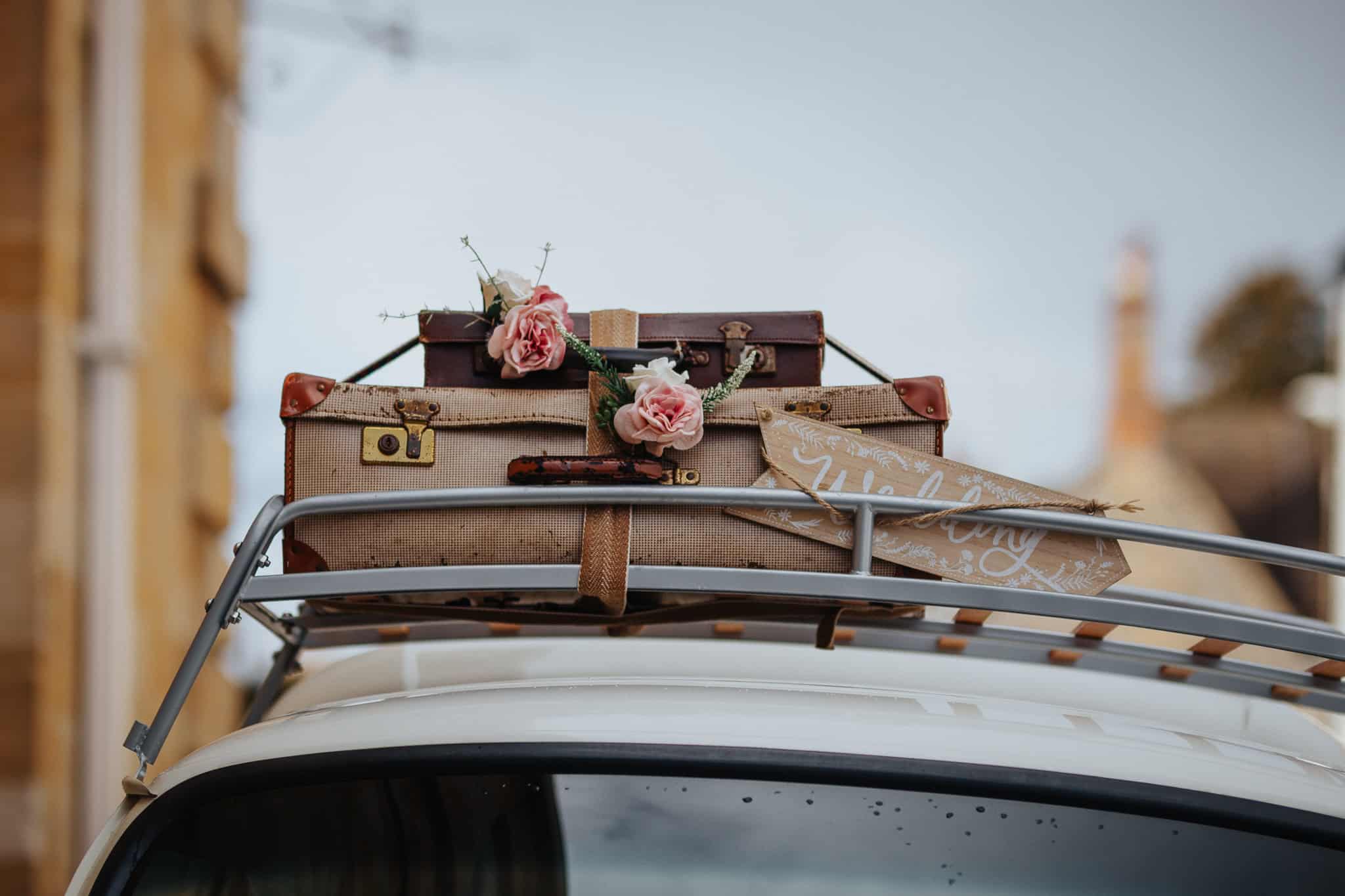 Close-up of a vintage VW wedding camper roof rack adorned with rustic suitcases, flowers, and a wooden "Wedding" sign. A charming and stylish wedding transport detail in Woodstock, Oxfordshire.