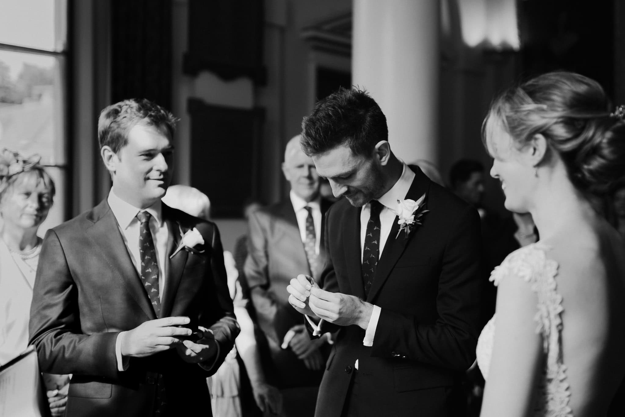 Black and white candid moment of the groom holding the wedding ring before exchanging vows at Woodstock Town Hall, with the bride and best man smiling. A heartfelt and joyful wedding scene.