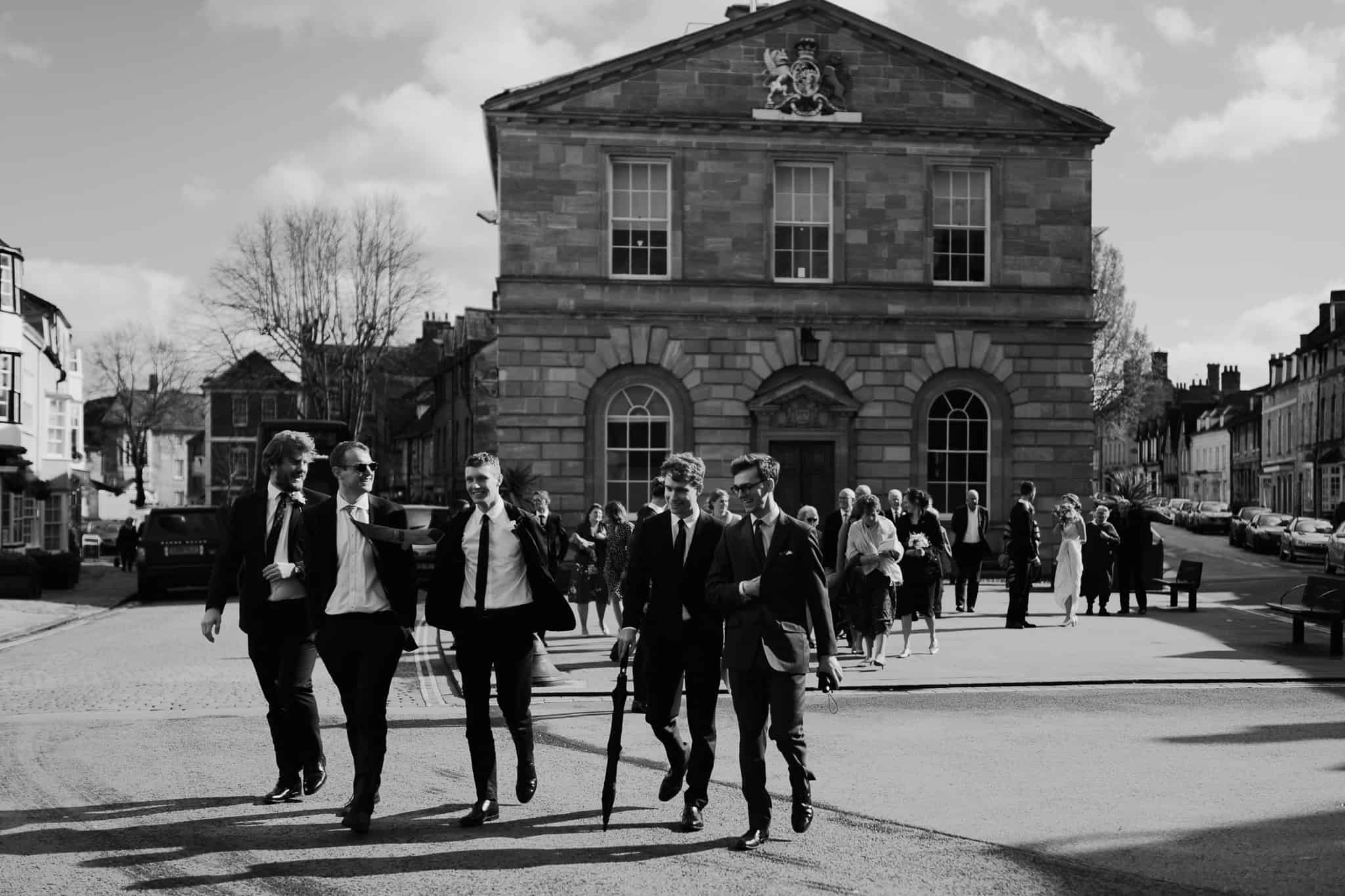 A joyful black and white candid of wedding guests laughing and walking outside Woodstock Town Hall, with the bride and groom following behind. A beautifully captured celebratory moment.