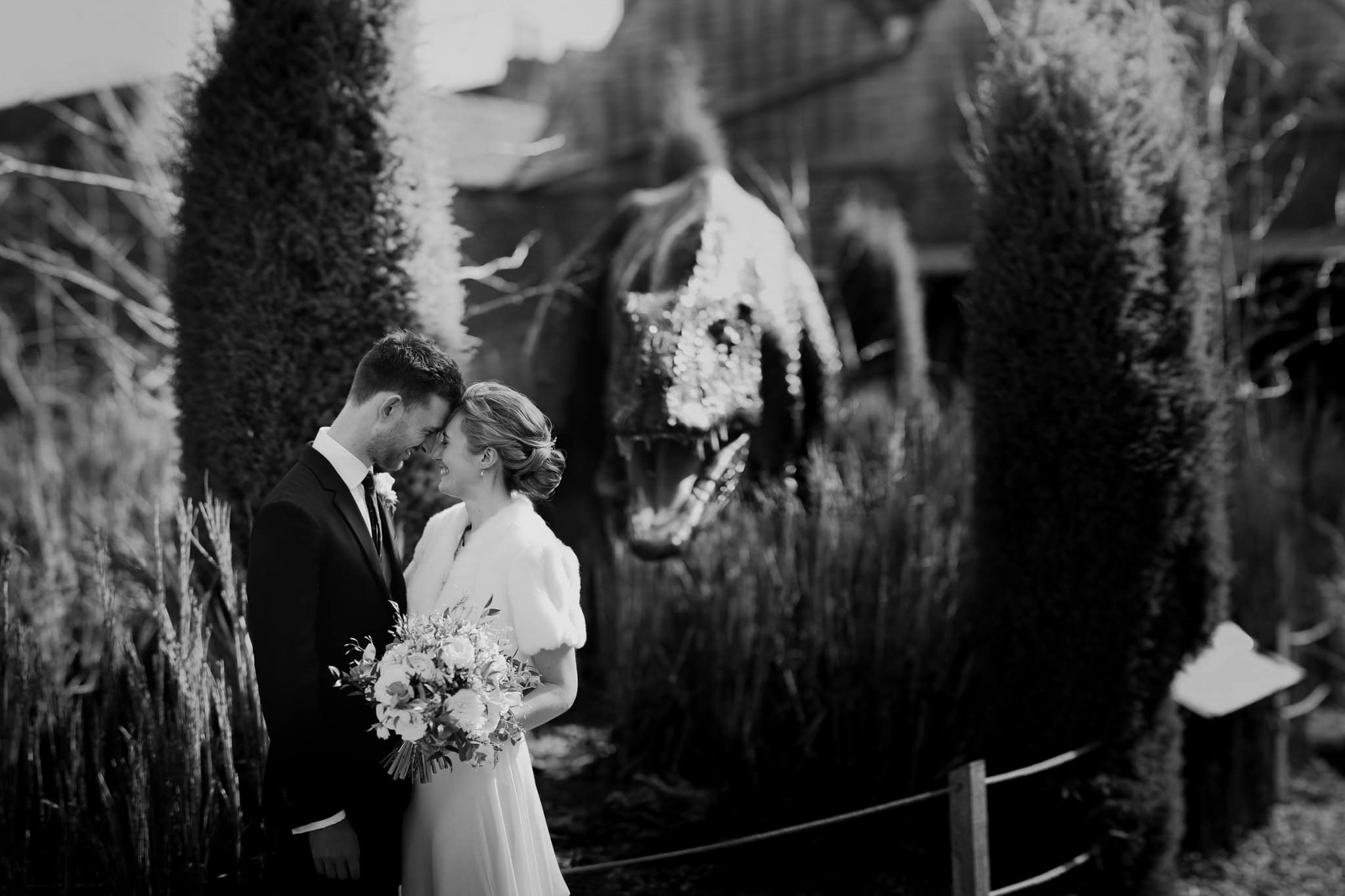 Bride and groom share a heartfelt moment, foreheads touching, with a dramatic dinosaur sculpture in the background. A unique and artistic wedding portrait captured in Oxfordshire.