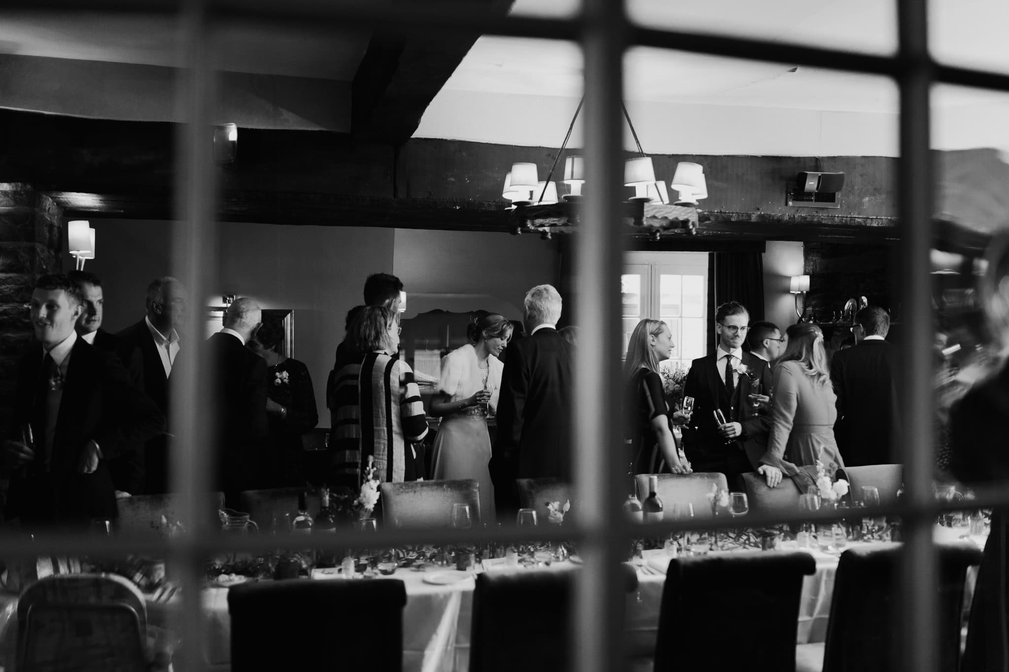 A candid black and white shot through the window capturing guests mingling at Alice and Kit’s wedding reception at Macdonald Bear Hotel in Woodstock, Oxfordshire. Elegant and timeless wedding photography.