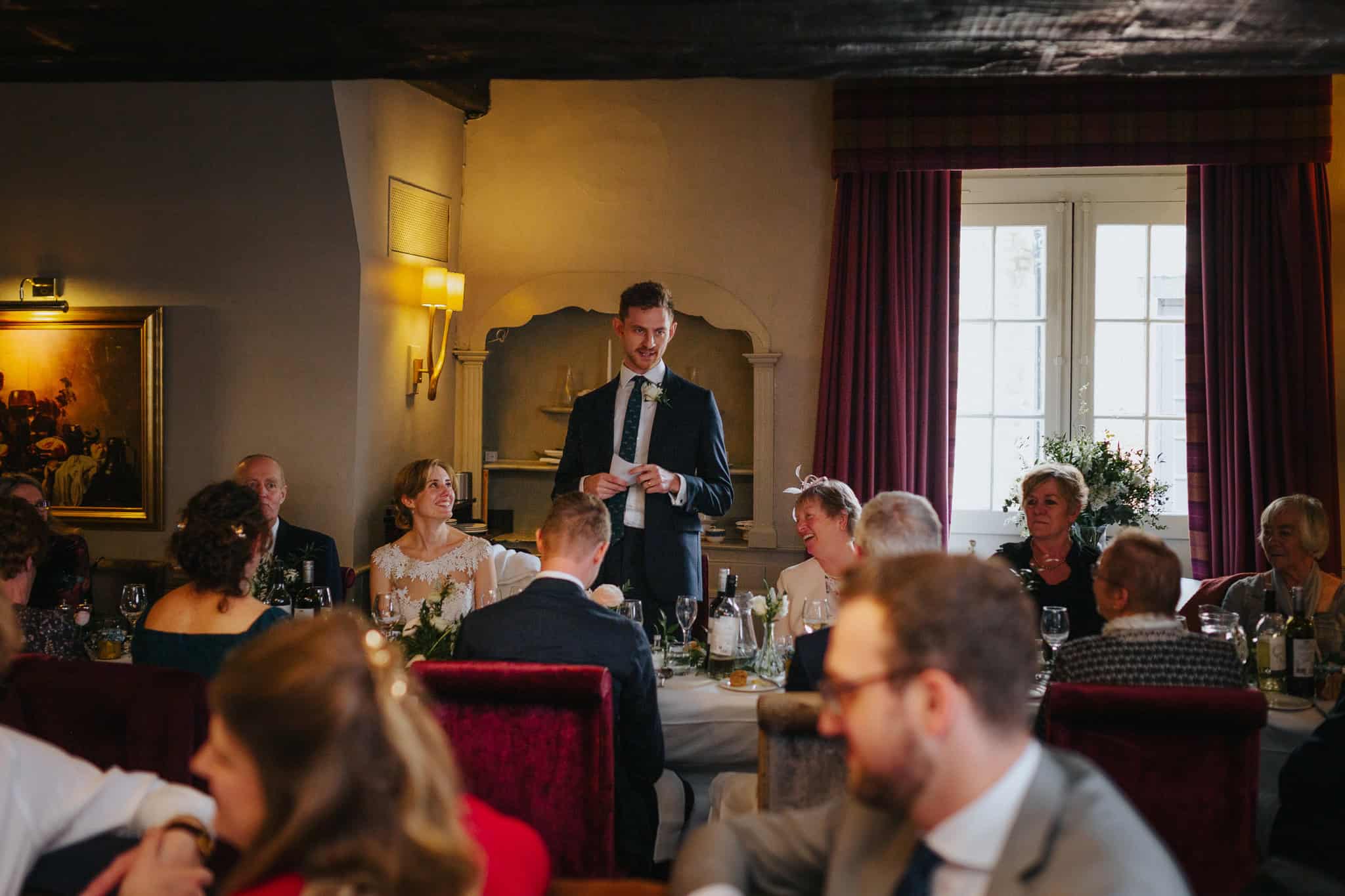 The groom delivers a heartfelt speech as the bride and guests smile at the wedding reception at Macdonald Bear Hotel, Woodstock. A candid moment capturing the joy of a wedding celebration.