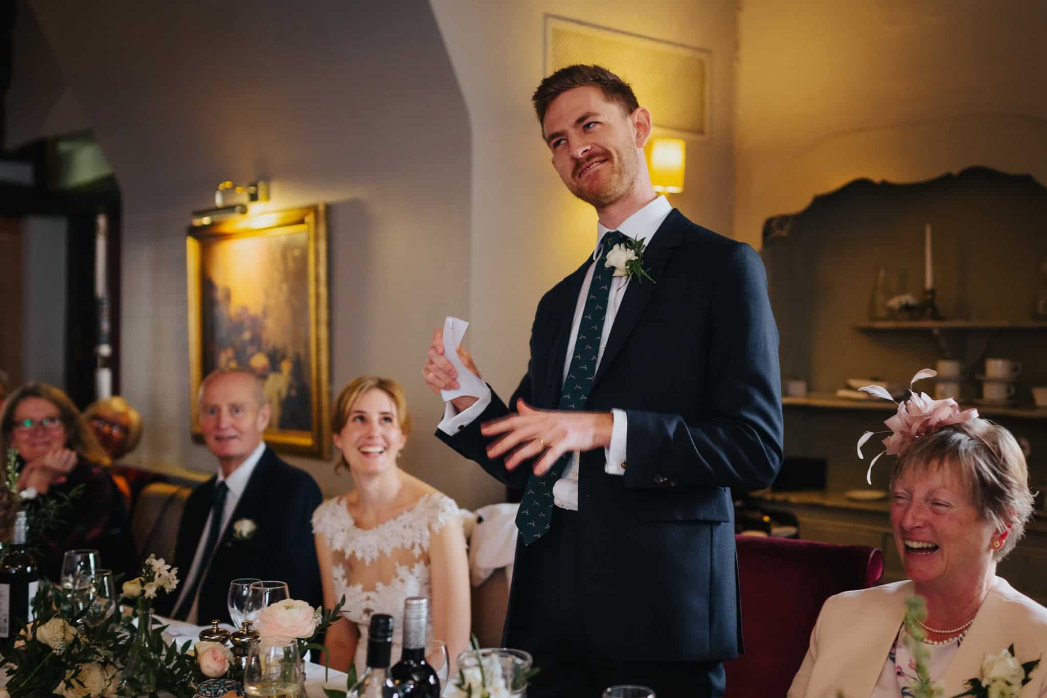 The groom delivers a witty and heartfelt speech at the wedding reception in Macdonald Bear Hotel, Woodstock, making guests, including the bride, laugh. A candid moment of joy and celebration.