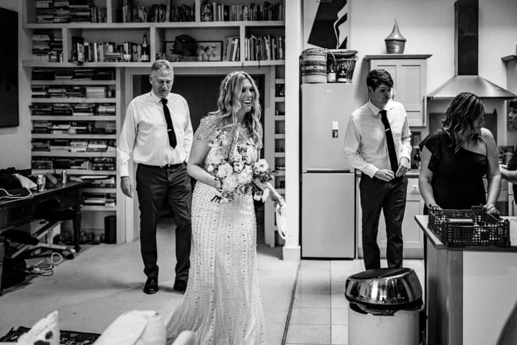 The photo captures a candid moment of wedding preparations in a London home. A bride, smiling joyfully while holding a bouquet of flowers, is surrounded by family and friends. In the background, a man in a white shirt and black tie walks toward her, while another person adjusts something on her dress. The scene is set in a cozy kitchen with bookshelves visible, giving it a warm, homey atmosphere. The bride’s sparkling dress contrasts with the casual attire of the others, adding to the lively, intimate vibe of the moment.
