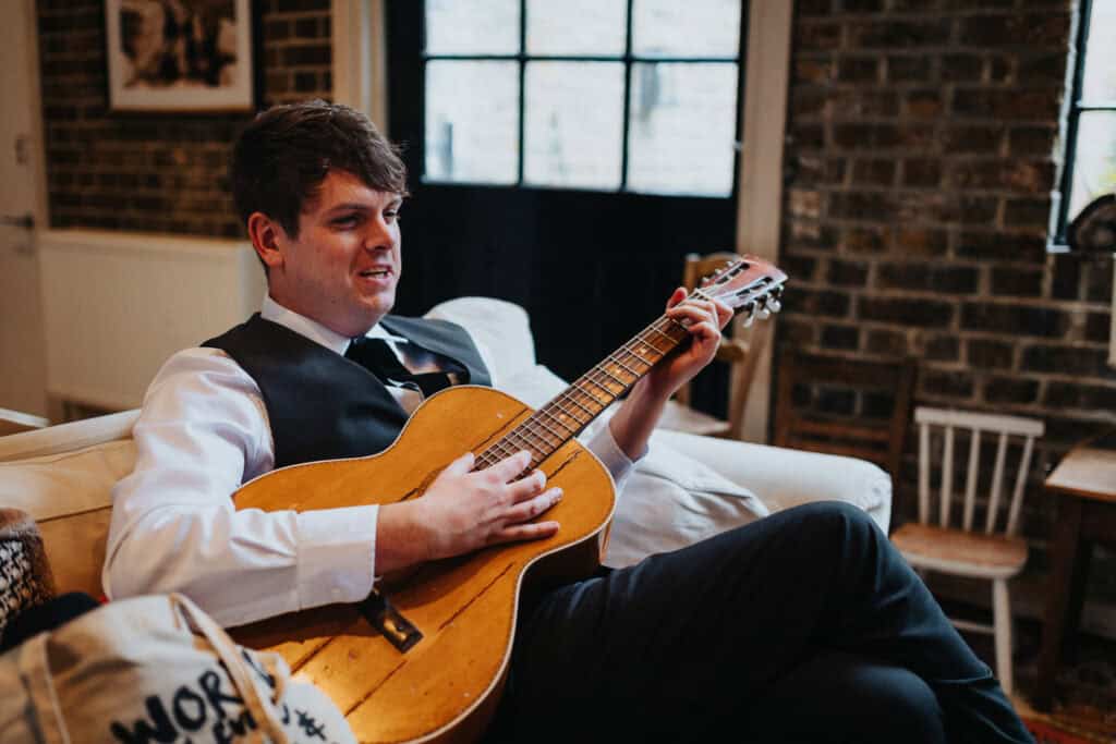 The brother of the bride, dressed in a white shirt and vest, sits on a sofa playing an acoustic guitar during wedding preparations at a charming venue in London. The brick wall and natural light from the windows create a warm, relaxed atmosphere.