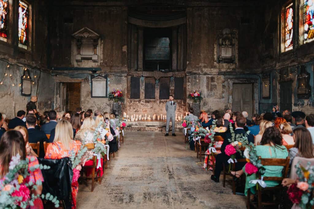 A wedding ceremony taking place in a historic asylum chapel in London. The groom, Tom, stands at the altar, waiting for his bride. The venue features a rustic, atmospheric setting with vintage stained-glass windows, exposed brick, and candles lining the floor. Guests are seated on wooden chairs, adorned with floral decorations, as they look toward the groom. The space has a mix of industrial and romantic vibes, with fairy lights adding a soft glow to the atmosphere.