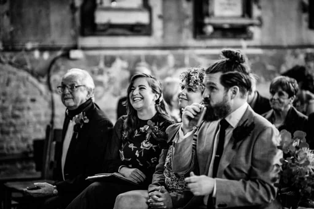 Guests enjoying the wedding ceremony at the Asylum Chapel in London, with a candid moment of laughter and conversation. The best man, seated in the front row, is seen holding a drink, dressed in a suit with a boutonnière, while other guests share the moment.