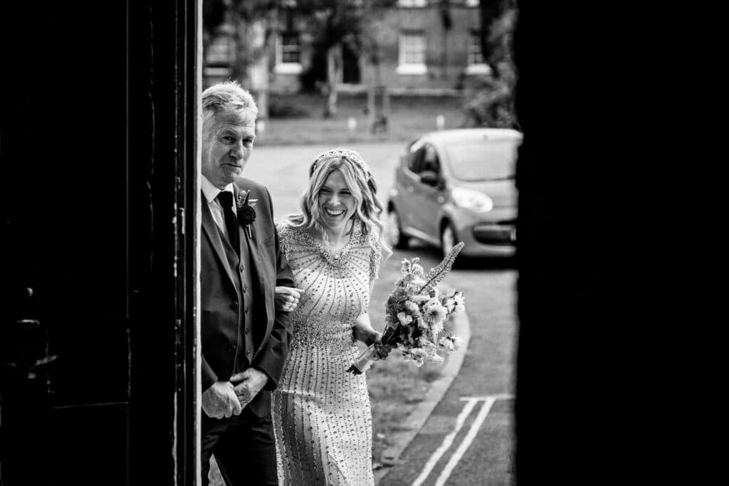 A bride in a beaded gown, holding a bouquet, smiles as she walks arm-in-arm with her father toward the ceremony. They are entering a wedding venue, with the entrance door framed in the foreground. The setting is a wedding ceremony at the Asylum Chapel in London