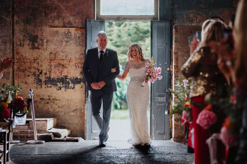 A bride in a sparkling, elegant dress smiles as she walks down the aisle with her father during a wedding ceremony at the Asylum Chapel in London. The rustic, charming setting features a mix of exposed brick walls and natural light streaming in from the open doors, creating a warm and joyous atmosphere.