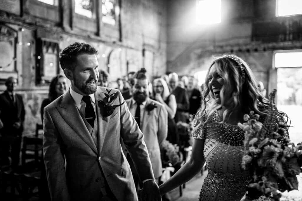 Aimee, the bride, and Tom, the groom, share a joyful moment during their wedding ceremony at an asylum chapel in London. The couple holds hands, smiling warmly at each other as they walk down the aisle. The chapel's vintage atmosphere and natural light enhance the romantic and intimate setting of their special day.