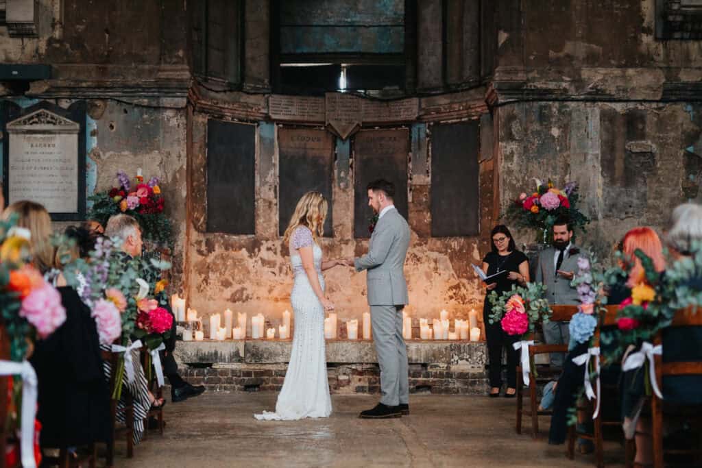 Wedding ceremony at the Asylum Chapel in London, with the couple exchanging wedding rings in front of a beautifully decorated altar. The bride, wearing a lace wedding dress, and the groom, dressed in a grey suit, are standing together as they exchange vows. The intimate setting features candles, vibrant flowers, and guests seated with floral wreaths adorning their chairs.