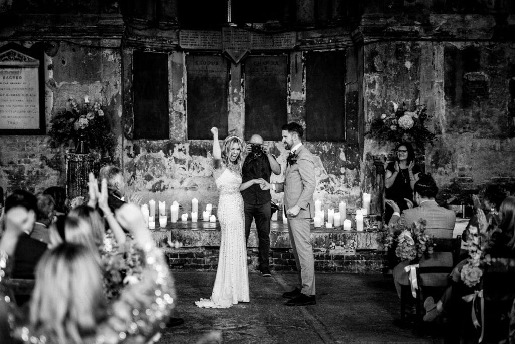 Bride joyfully raises her arm during a wedding ceremony in an abandoned chapel in London. The groom stands beside her, smiling, while guests applaud.