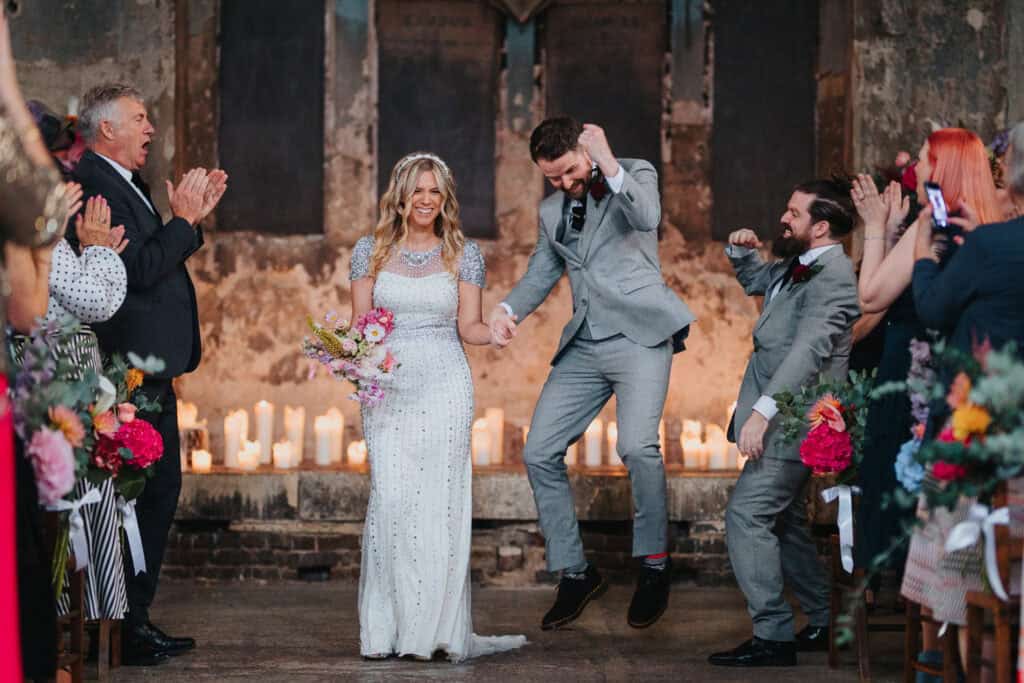 A joyful couple exits their wedding ceremony at an asylum chapel in London. The bride, wearing a sparkling white gown and holding a colorful bouquet, smiles as she holds hands with the groom, who is playfully jumping in excitement. Guests cheer and applaud in the background, with candles lining the chapel's stone walls, creating a warm and festive atmosphere.
