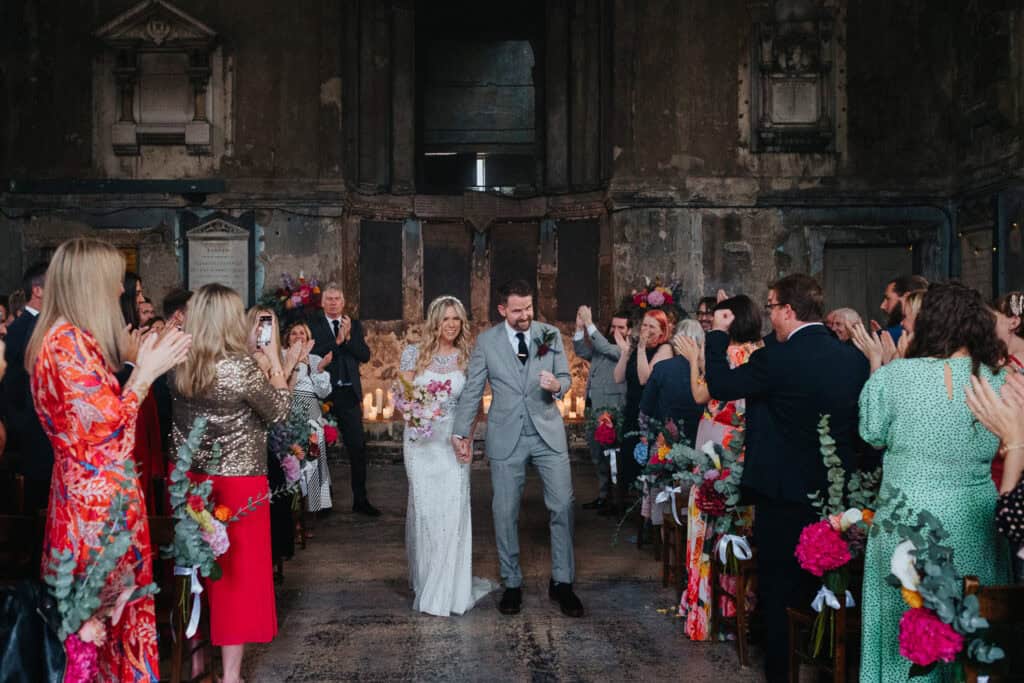 A joyful wedding ceremony taking place in the unique setting of an asylum chapel in London. The bride, wearing a sparkling white dress and holding a colorful bouquet, walks hand-in-hand with her groom, who is dressed in a gray suit. They are greeted by their applauding guests, surrounded by candles and vibrant floral arrangements. The ceremony has an intimate, vintage atmosphere with the chapel’s old, textured walls in the background.