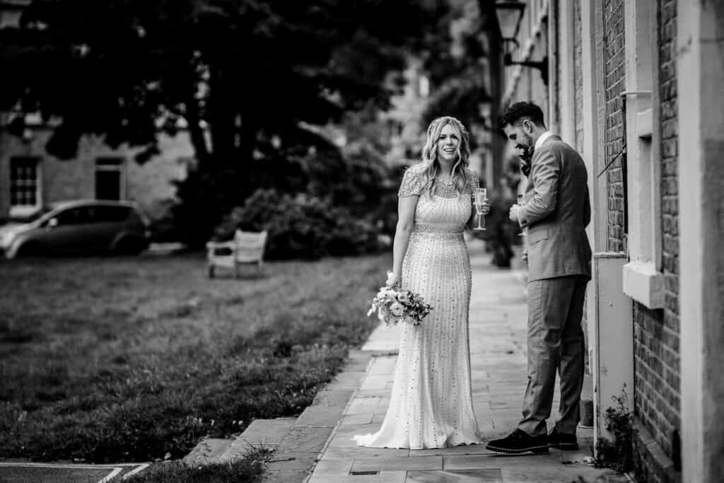 A quiet, intimate moment after the wedding ceremony at the Asylum Chapel in London. The bride, in a beautiful beaded gown, smiles softly while holding her bouquet. The groom, dressed in a suit, stands beside her, adjusting his attire. The photo, in black and white, captures a peaceful atmosphere before the confetti photo, with the chapel's architecture subtly in the background.