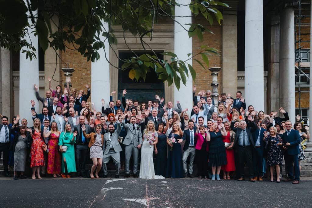 A large group of wedding guests gathered outside the Asylum Chapel in London, posing for a photo. The guests are dressed in formal attire, with some in bright colors, while the bride and groom stand at the front, smiling and waving. The scene captures a joyful celebration with a historic building in the background, featuring grand columns and a few trees framing the shot.