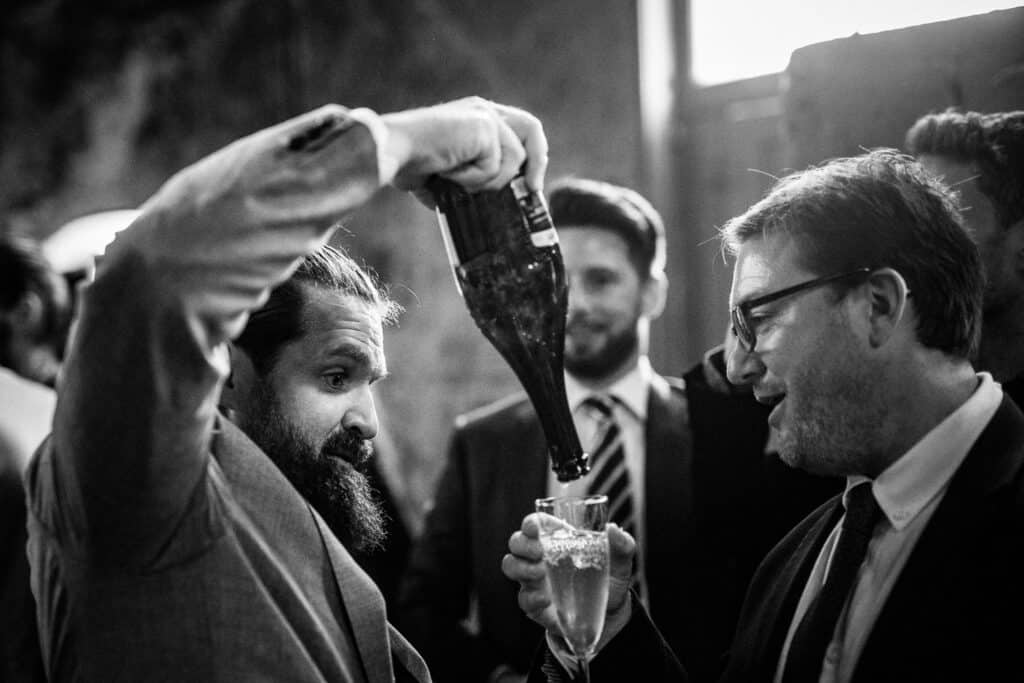 Two men celebrate with champagne at a wedding reception at the Asylum Chapel in London. One man, with a beard, pours champagne into the other's glass. Both are dressed in formal attire, with the man holding the bottle smiling while the other man raises his glass in appreciation. The background features other wedding guests and the unique architecture of the chapel.