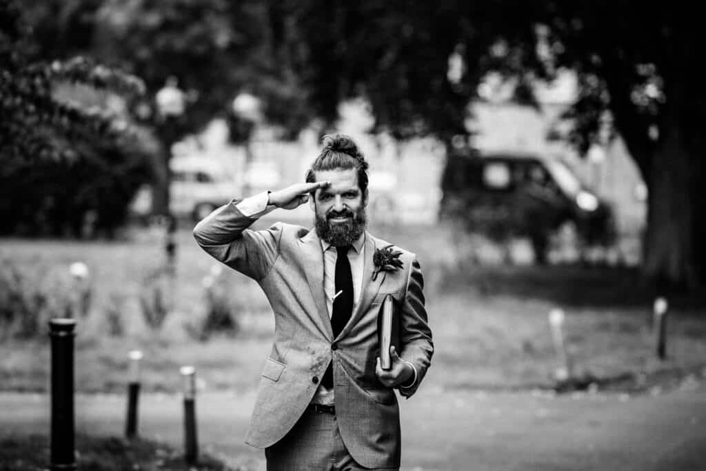 A stylish best man saluting with a smile, wearing a grey suit, black tie, and a boutonnière, while holding a folder in one hand. The photo is taken in black and white, with a blurred background of a park setting near the Asylum Chapel in London, capturing a cheerful and confident moment.
