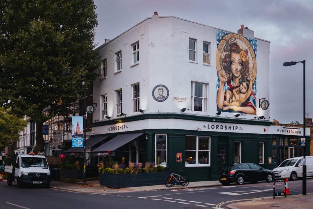 A lively scene from the wedding celebration of Aimee and Tom at The Lordship Pub, showcasing the venue's distinctive corner building with a striking mural on the exterior wall. The vibrant street scene includes trees, parked vehicles, and the pub's welcoming entrance with 'Pub & Kitchen' signage, creating a warm and festive atmosphere for the event.