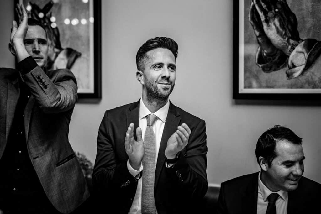 The photo captures a moment during the wedding celebrations of Aimee and Tom at The Lordship Pub. It shows several guests applauding, with one man in a suit smiling and clapping, expressing joy and excitement. The atmosphere appears lively and celebratory, reflecting the warmth and happiness of the occasion.