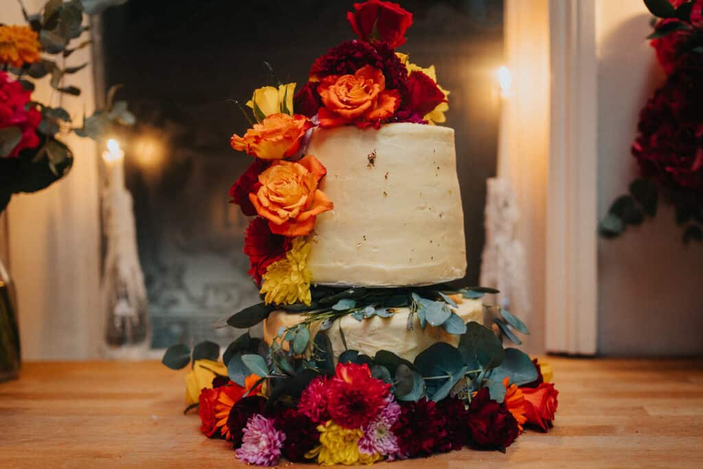 Wedding cake at the celebration of Aimee and Tom at The Lordship Pub. The cake features a rustic, layered design adorned with vibrant orange, red, yellow, and purple flowers, as well as lush greenery, creating a warm and colorful centerpiece for the event.