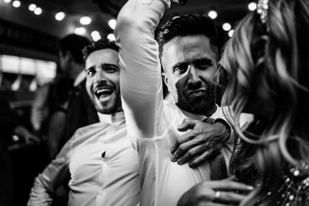 A lively moment from Aimee and Tom's wedding celebrations at The Lordship Pub, capturing guests dancing and enjoying the festivities. Two men, one with a big smile and the other with a playful expression, are in the midst of the celebration, with a third woman dancing nearby. The atmosphere is vibrant and joyful, illuminated by warm string lights overhead.