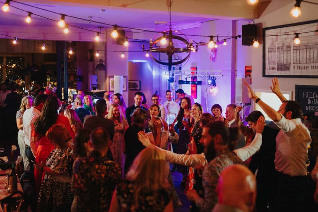 Guests dancing and enjoying the evening party at The Lordship Pub during Aimee and Tom’s wedding. The lively atmosphere is highlighted by string lights, colorful lighting, and happy, engaged guests celebrating together on the dance floor.