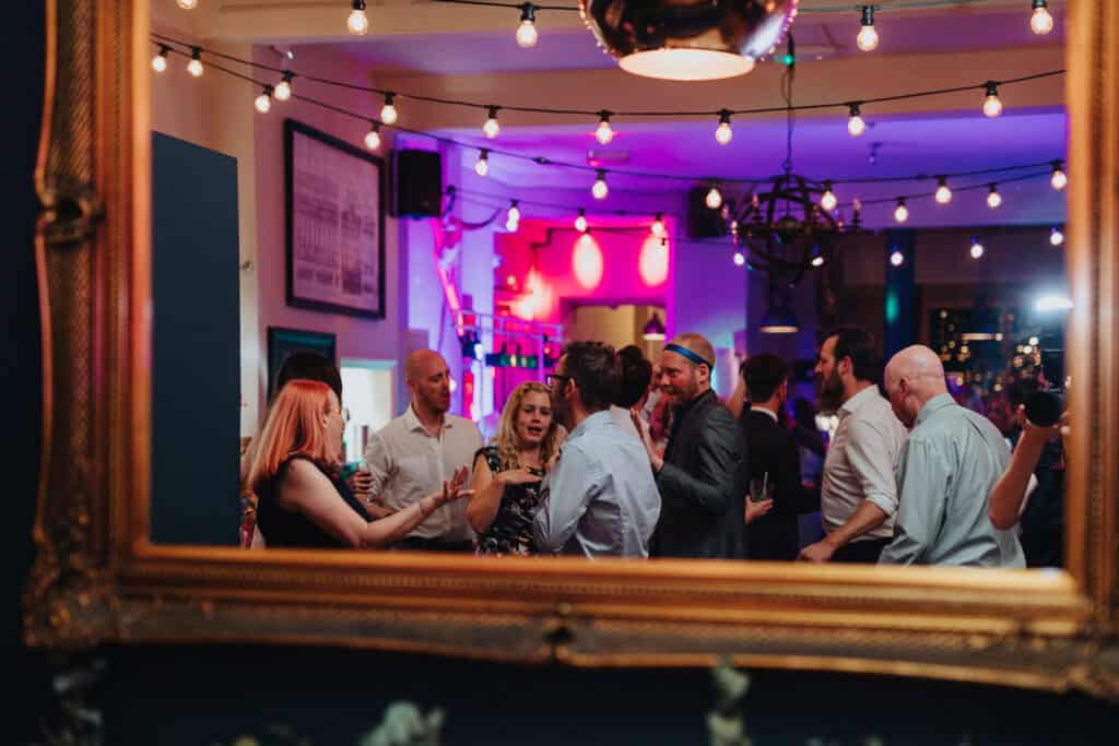 The photo shows a lively evening party at The Lordship Pub during the wedding of Aimee and Tom. The scene is captured through a mirror, with guests enjoying themselves, chatting, and dancing in a vibrant atmosphere. The venue is decorated with string lights and a mix of colourful lights, creating a fun and festive vibe. People are dressed in formal attire, and there’s a mix of animated conversations and laughter as the celebration continues.
