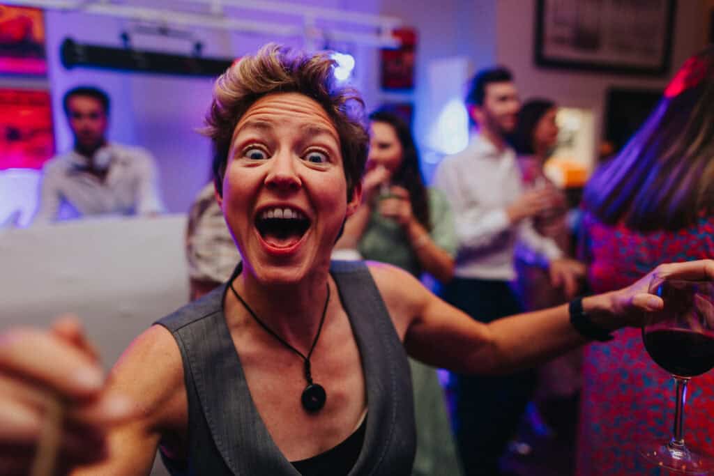 A lively moment from the evening party at The Lordship Pub during Aimee and Tom’s wedding. A woman with short, tousled hair, wearing a gray dress and a necklace, is enthusiastically dancing and laughing. She is holding a glass of red wine, surrounded by other guests enjoying the celebration. Colourful lights and a festive atmosphere complete the scene.