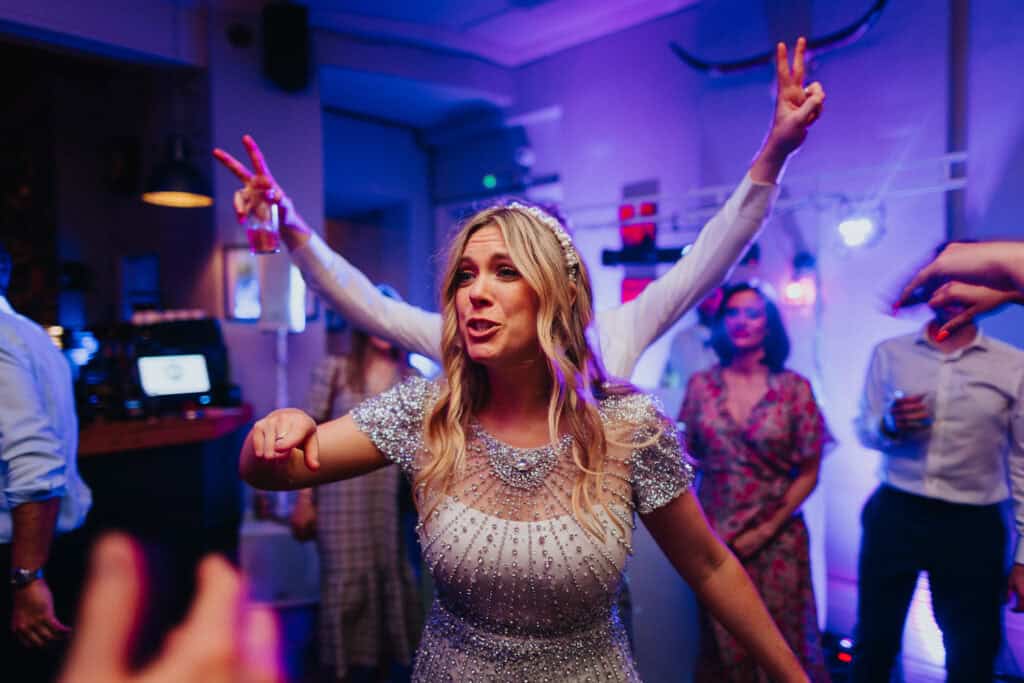 Aimee enjoying the evening party at The Lordship Pub during her wedding reception with Tom. She is dancing and laughing, wearing a sparkling, beaded dress and a pearl headband. The colourful lighting creates a lively atmosphere as guests celebrate.