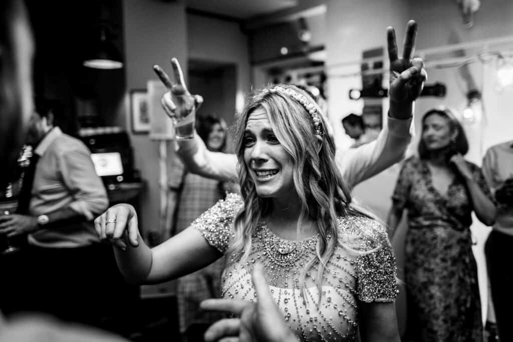 Bride Aimee at her evening wedding party at The Lordship Pub, enjoying the celebration. She is smiling, with her hand raised holding a drink, surrounded by guests in a lively atmosphere. The image captures a candid moment of joy, with people dancing and celebrating in the background.