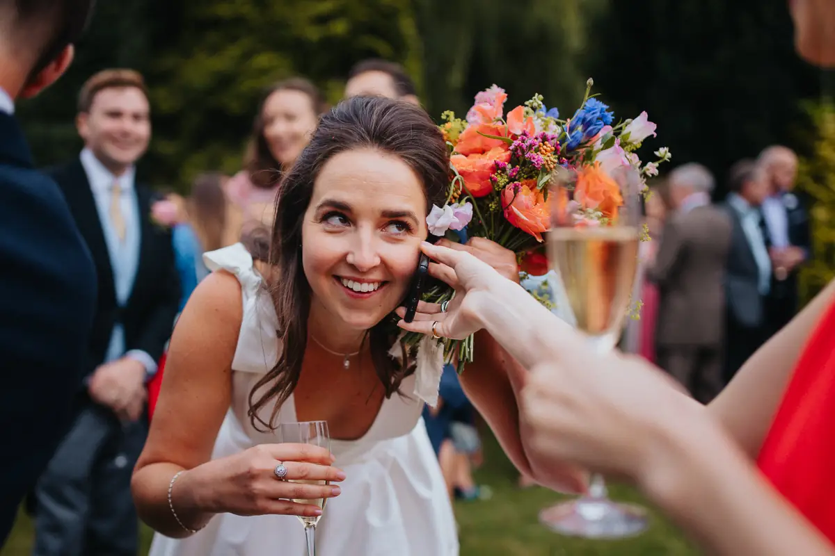 A vibrant moment as the bride smiles, holding a glass of champagne and a colourful bouquet, while answering a call during the celebration. Full of personality and energy.