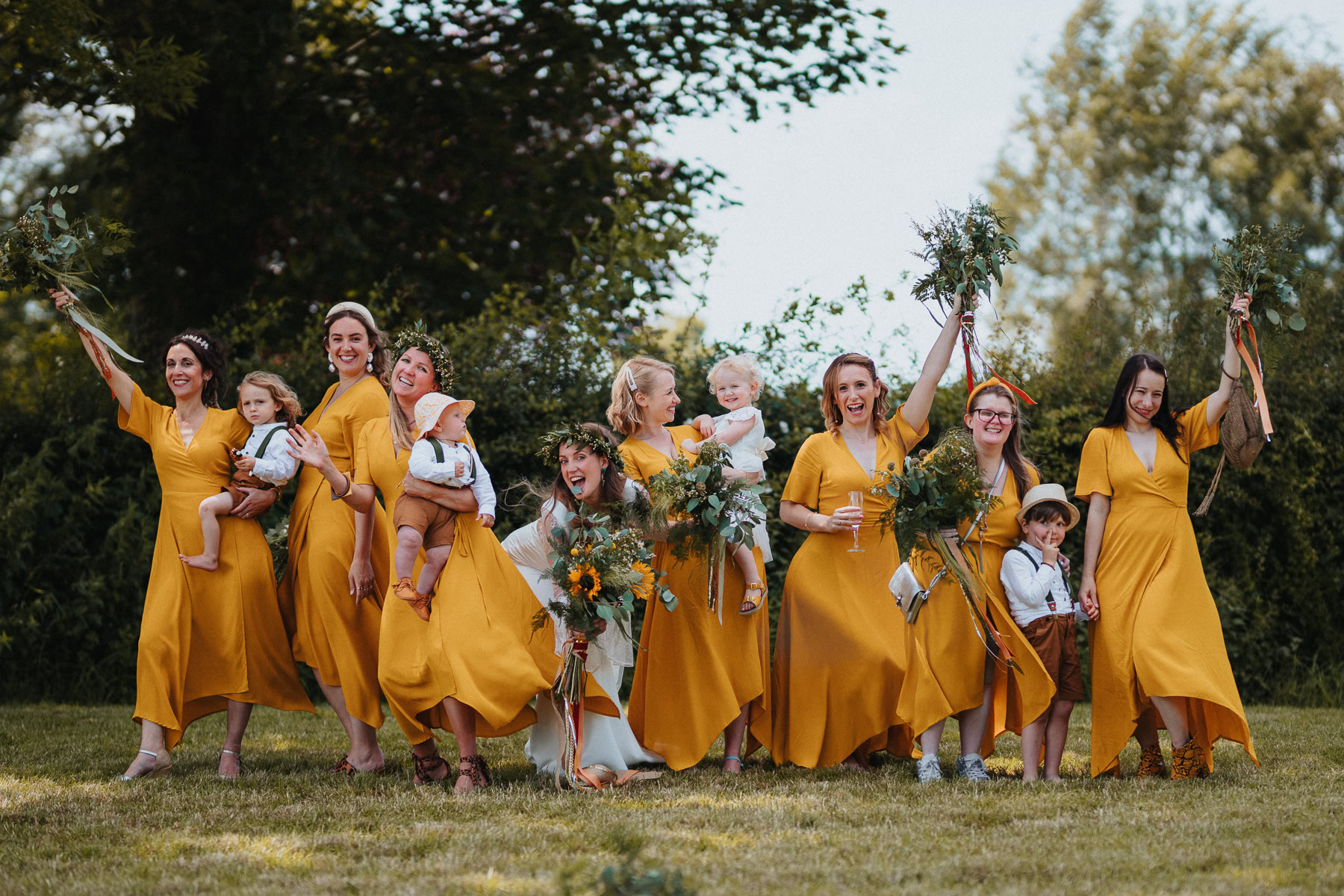 A joyful group of bridesmaids dressed in mustard yellow dresses celebrating at a farm wedding, holding bouquets of greenery and flowers. Some are carrying young children dressed in coordinating outfits, while others cheerfully raise their bouquets. The natural, outdoor setting with lush greenery in the background enhances the warm and rustic wedding theme.