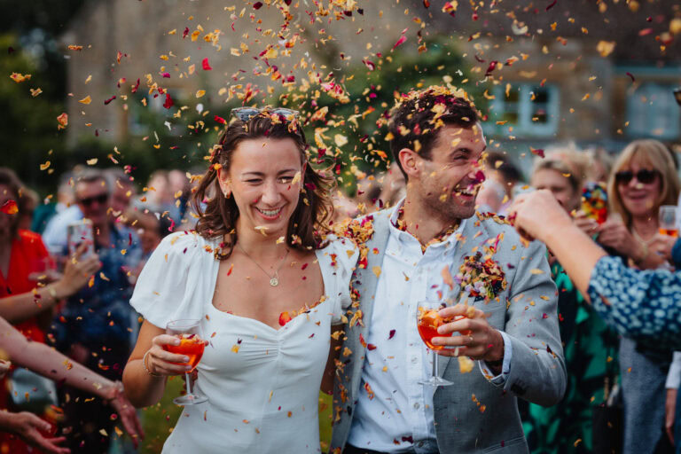A newlywed couple walks through a joyful confetti shower, smiling and laughing as they hold Aperol Spritz drinks. The bride wears a white dress with puffed sleeves, and the groom sports a light grey suit with a boutonnière. Surrounded by cheering guests in colorful attire, the moment is full of energy, celebration, and love. Captured outdoors with a blurred countryside setting in the background.