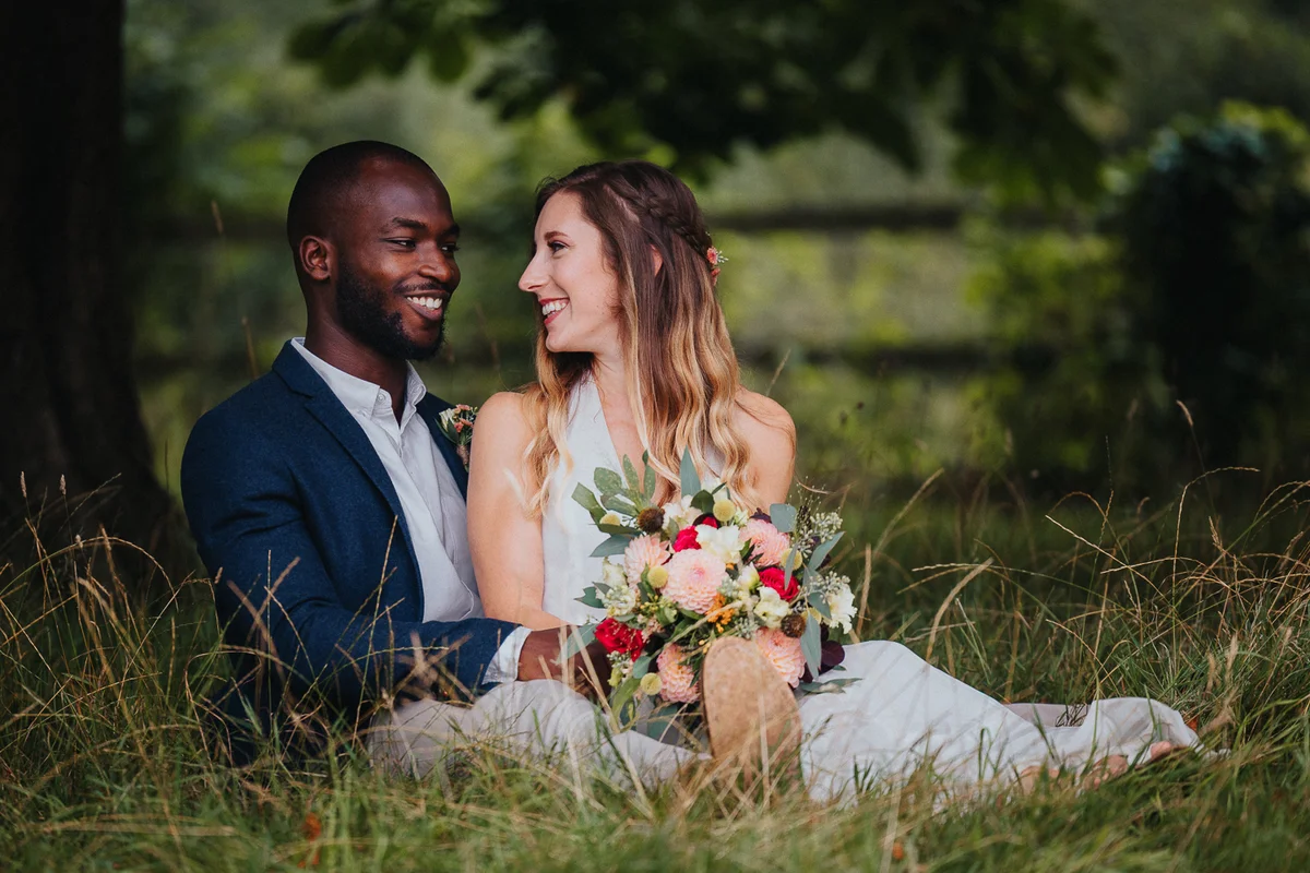 Bride and groom sitting on the grass, smiling at each other with bouquet
