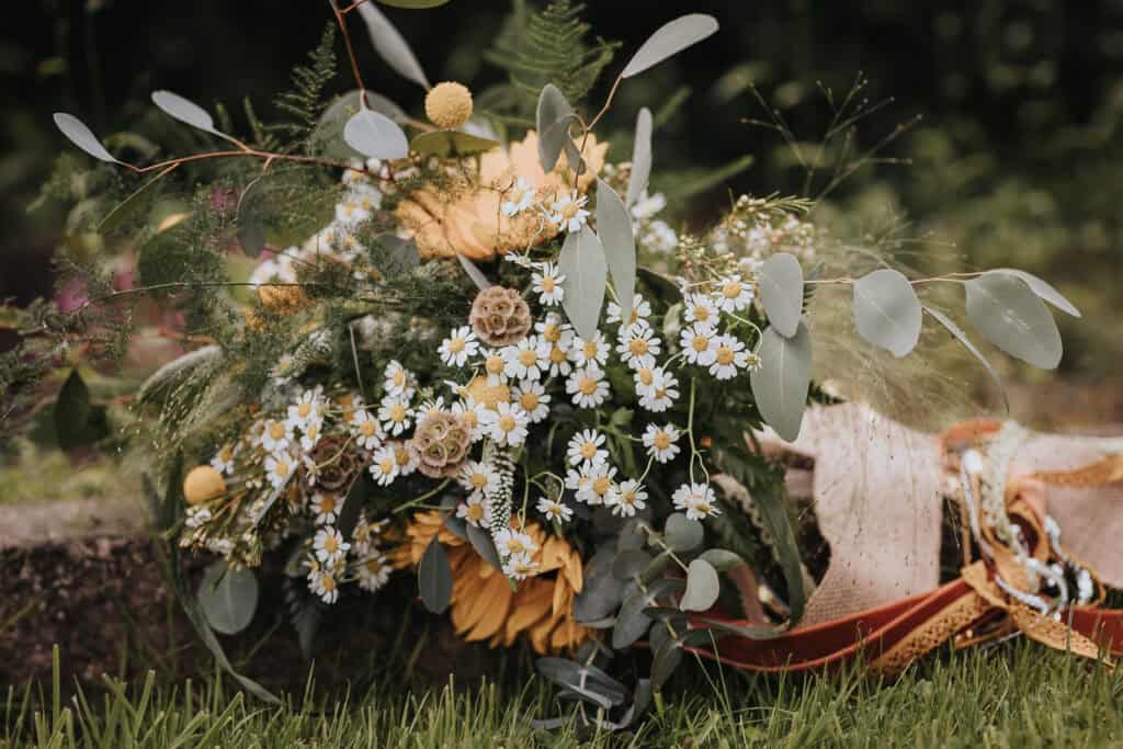 Rustic wildflower bouquet featuring sunflowers, daisies, eucalyptus leaves, and natural accents, styled with an organic and romantic aesthetic, displayed outdoors on lush green grass.