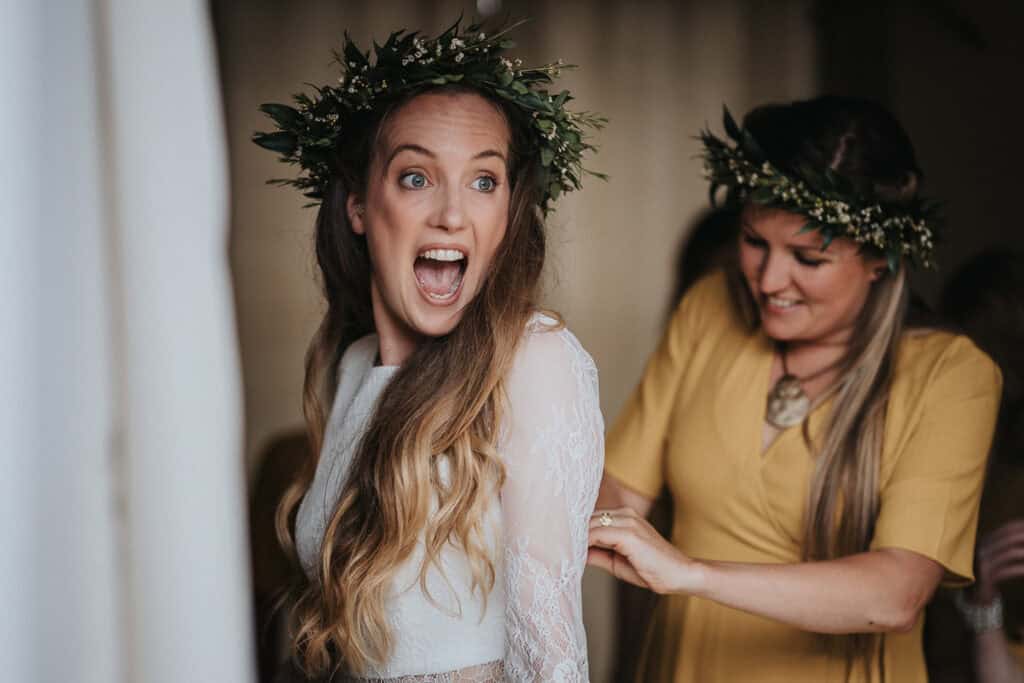 Joyful bride getting ready for her farm marquee summer wedding, with her bridesmaid assisting her, both adorned with rustic floral crowns.