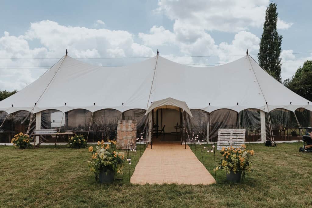 Large white wedding marquee set up on a grassy lawn, featuring a paved walkway leading to the entrance, surrounded by flower arrangements and rustic wooden furniture under a partly cloudy sky.