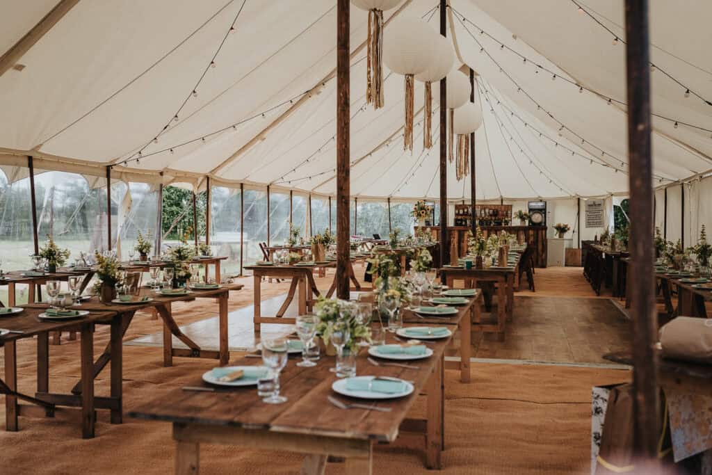 A rustic wedding marquee with wooden tables set for a reception. Tables are adorned with elegant floral centrepieces, light blue plates, and glassware, under a canopy of string lights and hanging lanterns. The setting combines natural wooden decor and open views of the surrounding greenery, creating a charming and inviting atmosphere.