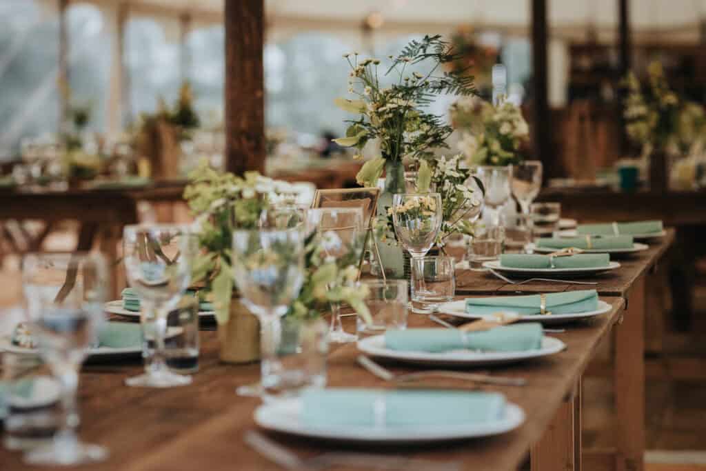 Rustic wedding table setup featuring a long wooden table adorned with elegant glassware, white plates, and teal napkins. Greenery and floral centrepieces add a natural, charming touch, set under a bright, airy marquee with warm natural lighting.