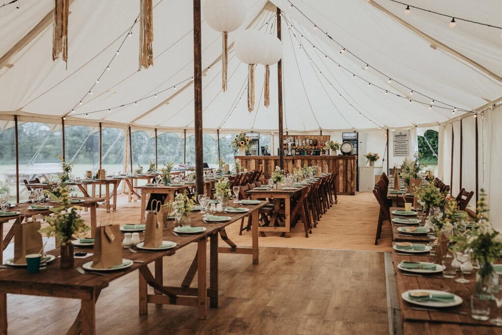 A beautifully decorated rustic wedding marquee featuring wooden tables with elegant floral centrepieces, green plates, and paper gift bags, complemented by string lights and a wooden bar at the far end. The marquee has a bright, airy ambiance with a natural wooden floor and large open windows.
