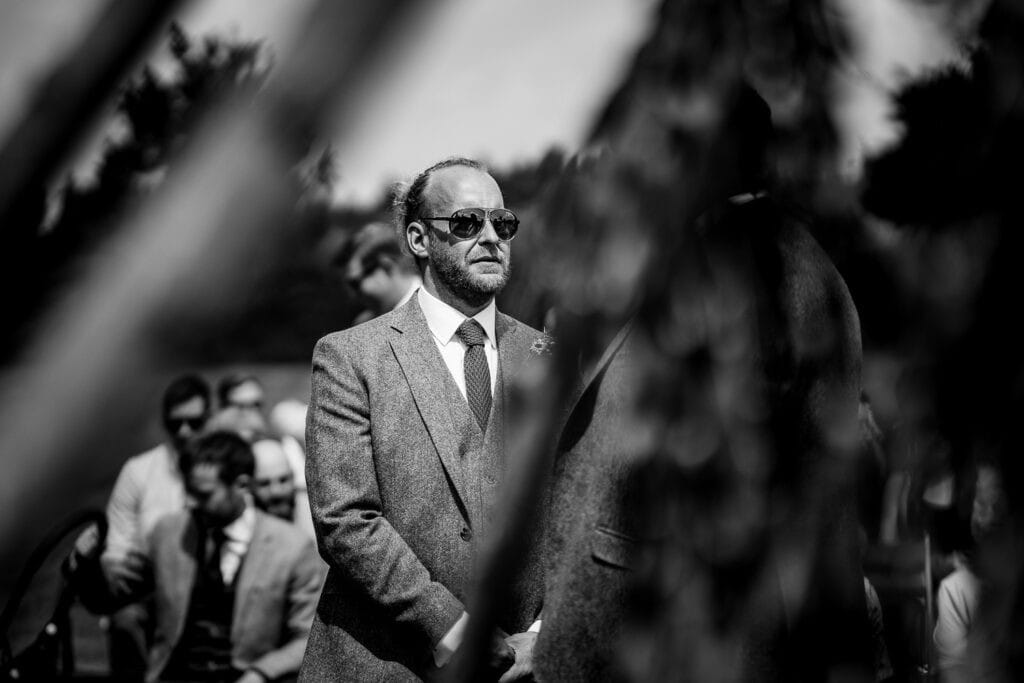 Black and white photograph from a farm marquee summer wedding, featuring the groom, Jon, wearing a suit and sunglasses, standing outdoors with blurred natural elements and guests in the background.