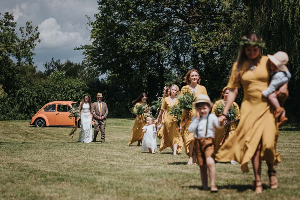 Capture the charm of an outdoor marquee wedding with this heartfelt moment: the bride walking arm-in-arm with her father, surrounded by her bridesmaids dressed in elegant mustard-yellow dresses, carrying lush green bouquets. Set against a picturesque backdrop of a vintage orange Beetle, vibrant greenery, and open skies, this scene embodies the perfect blend of rustic elegance and timeless celebration.