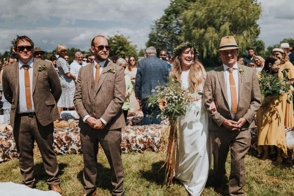 Celebrate love in a picturesque outdoor setting! This stunning marquee wedding captures the bride, radiantly adorned in an elegant white gown, walking arm in arm with her father. The natural beauty of the countryside serves as the perfect backdrop, with guests gathered to witness this heartfelt moment. The vibrant atmosphere, complemented by the stylish groom and groomsmen in classic tweed suits and earthy tones, showcases the charm of a relaxed yet sophisticated wedding celebration. Perfect for couples seeking a rustic and intimate outdoor experience.