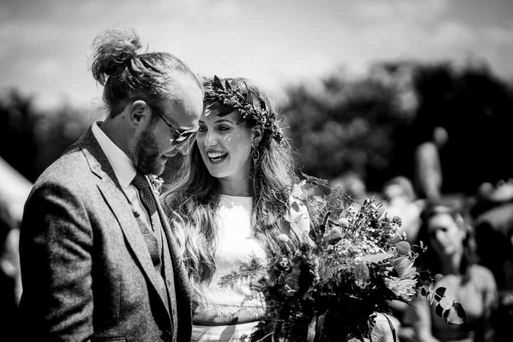 Black and white wedding photo of a joyful bride wearing a floral crown, holding a bouquet of wildflowers, smiling warmly at the groom dressed in a tweed suit and sunglasses, with an outdoor ceremony and blurred guests in the background.