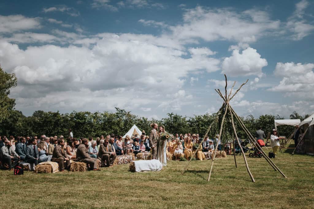 Outdoor rustic wedding ceremony in a picturesque countryside setting, featuring a large open field with wooden and hay bale seating, a decorative teepee structure, and a marquee tent in the background under a bright, partly cloudy sky.