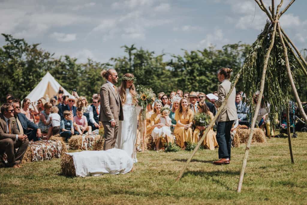 Outdoor marquee wedding ceremony set in a picturesque field, featuring a sunny summer sky, rustic hay bale seating, and a beautifully decorated natural altar surrounded by joyful guests.