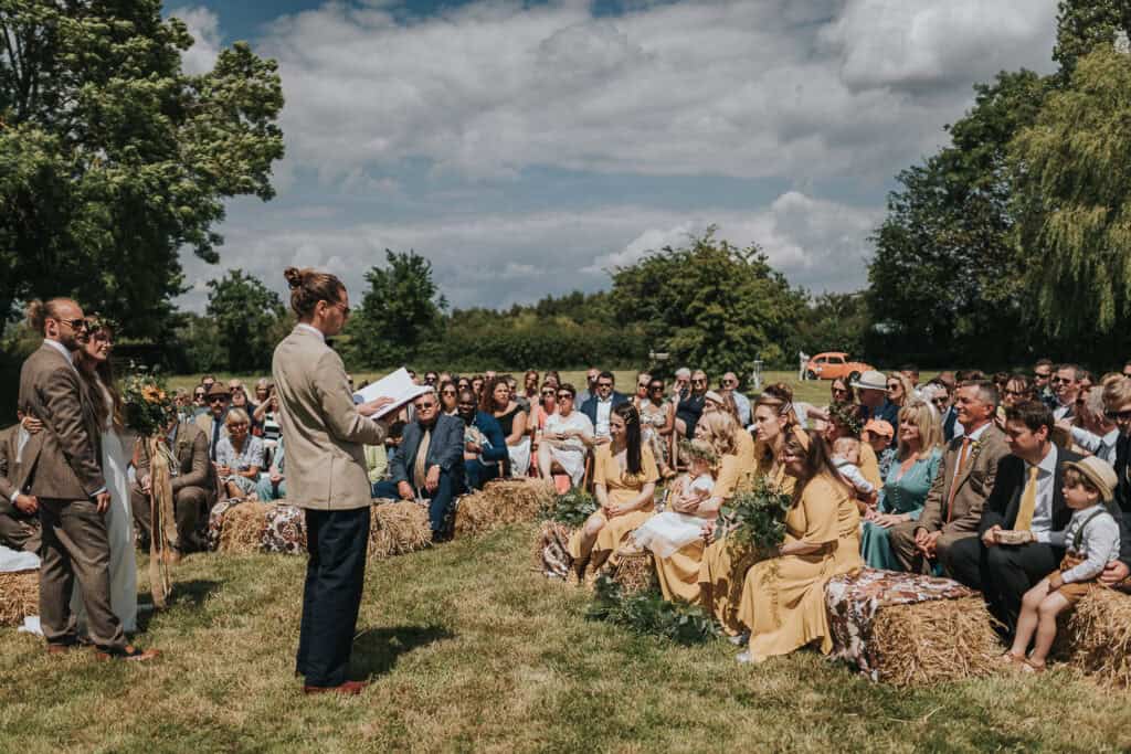 Outdoor summer wedding ceremony at a farm marquee venue, featuring guests seated on hay bales with vibrant greenery and clear skies in the background. The officiant, dressed in a beige jacket, is addressing the crowd, with the bridal party dressed in coordinated yellow outfits.